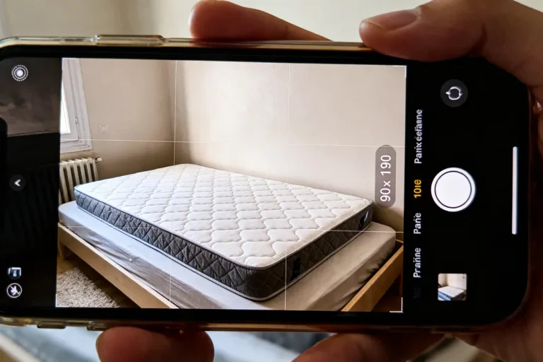 Smartphone photographiant un matelas sur un lit, avec écran de l'appareil photo affichant dimensions.