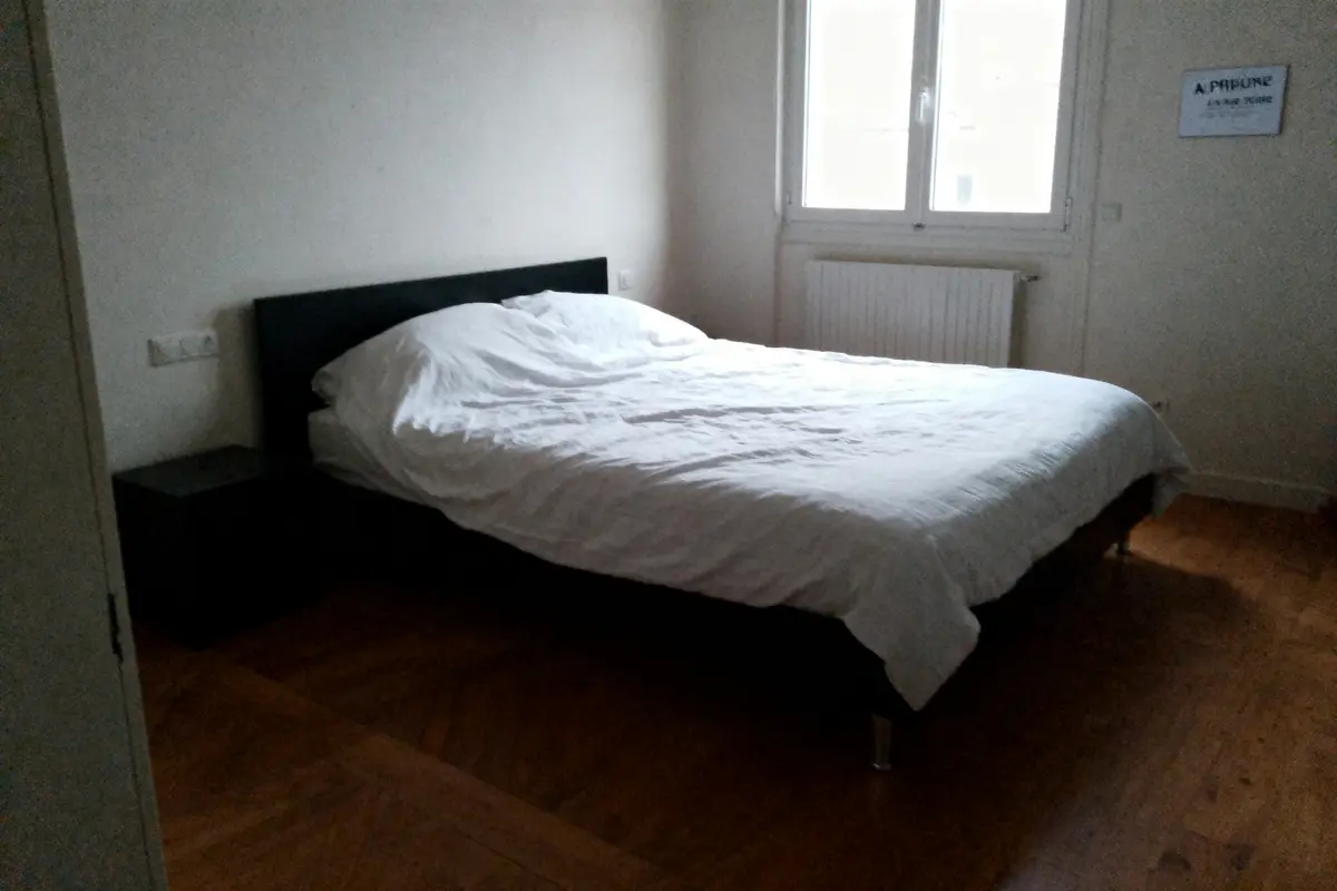 Lit double avec draps blancs, chambre lumineuse, parquet, fenêtre, table de chevet noire, murs clairs.